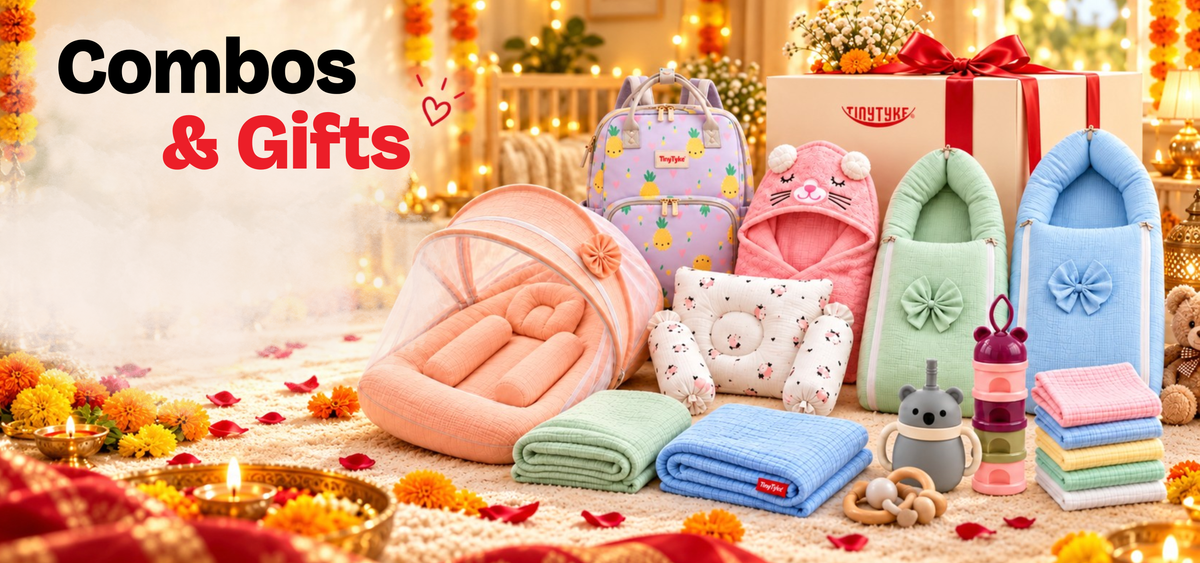 A baby gift set arranged on a soft blanket in a bright nursery-like setting. The set includes folded blankets, a pink bassinet, a patterned backpack, a bunny hood, a blue bassinet, folded towels, a bottle, and a large gift box tied with a red ribbon.